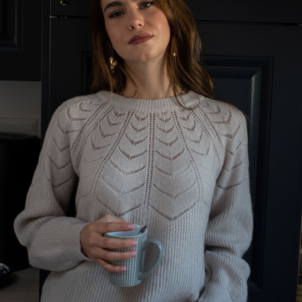 femme tenant une tasse de café dans la cuisine et portant un pull maille d'hiver écru avec détails ajourés